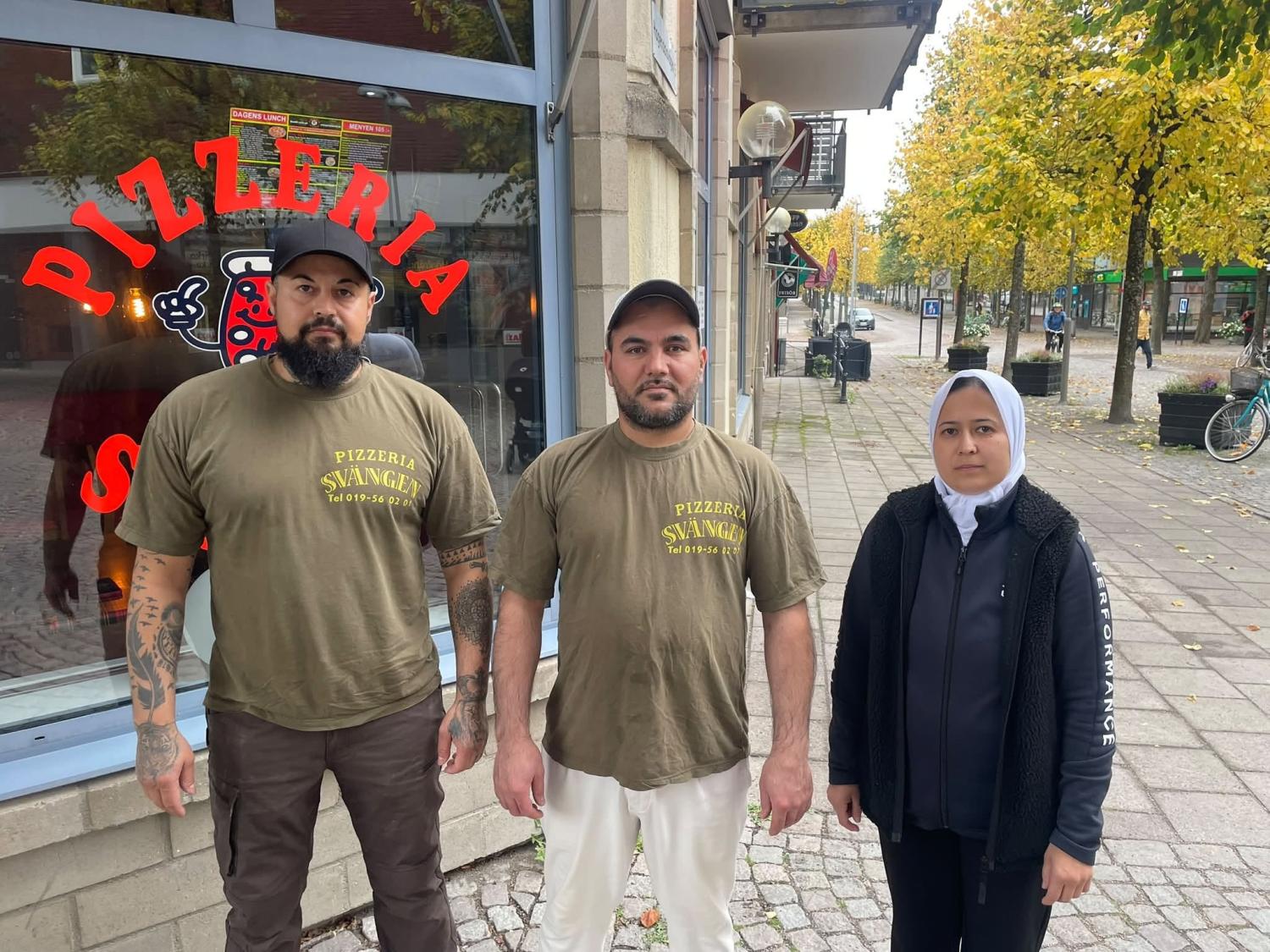 Tre personer står utanför en pizzeria på en trottoar av tegel, två av dem har på sig likadana olivgröna t-tröjor och en har en jacka med huva; träd med gula löv kantar gatan.