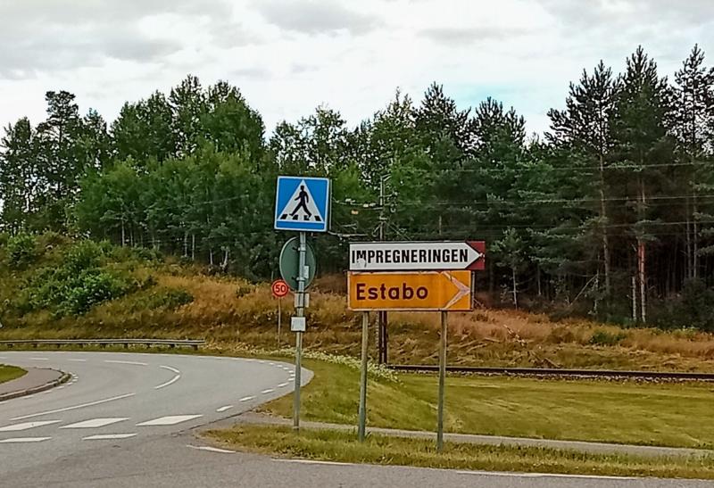 Vägkorsning med vägskyltar mot ”Impregneringen” och ”Estabo”, en skylt som anger övergångsställe samt träd i bakgrunden.