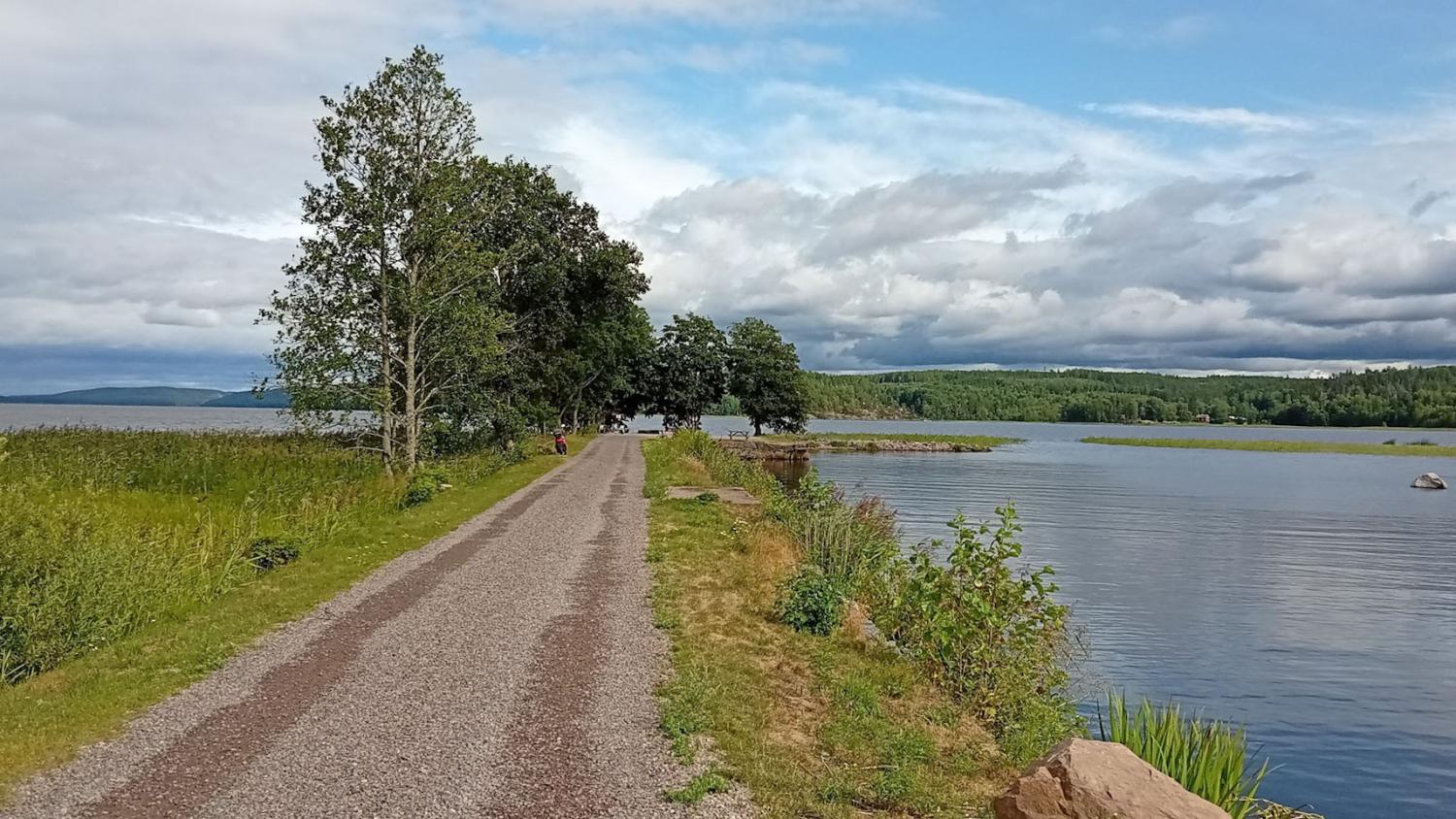 En grusväg löper längs en stilla sjö, omgiven av gräs och träd, under en delvis molnig himmel. I fjärran syns kullar och träd på andra sidan vattnet.