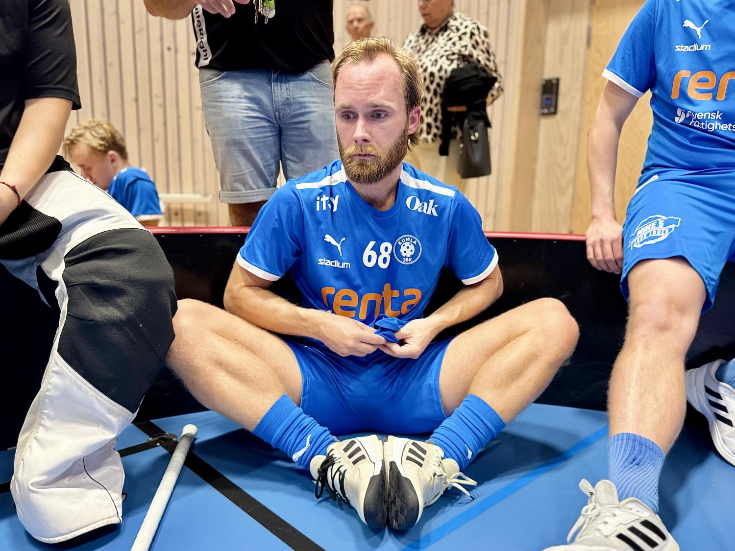 En innebandyspelare i blå uniform sitter på golvet med benen i kors och ser fundersam ut, omgiven av lagkamrater och utrustning.