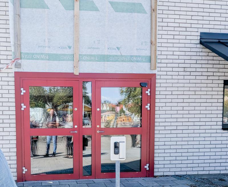 Röda dubbeldörrar med glasrutor inbyggda i en vit tegelvägg, med byggmaterial ovanför och en handdesinfektionsdispenser framför. I glaset syns speglingar av människor och träd.