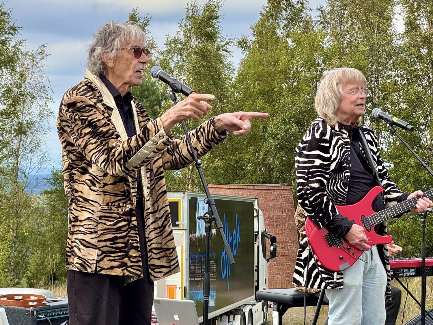 Två äldre män spelar musik utomhus; den ene sjunger i mikrofon och gestikulerar medan den andre spelar på en röd elgitarr. Båda bär jackor med djurmönster. Träd och utrustning syns i bakgrunden.
