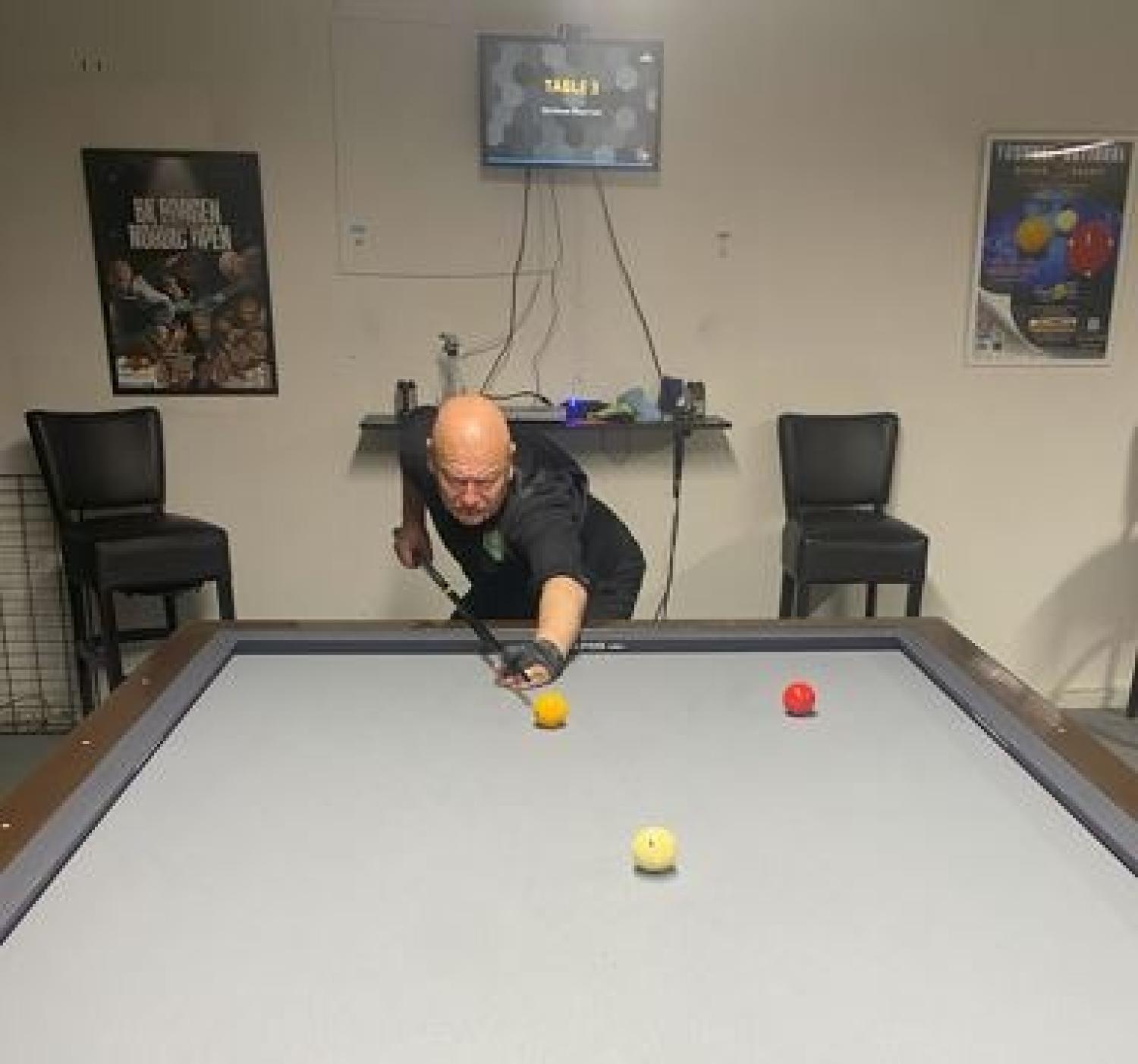 En man gör sig redo för ett skott på ett grått biljardbord med två gula och ett rött klot, i ett rum med affischer och stolar mot väggen.