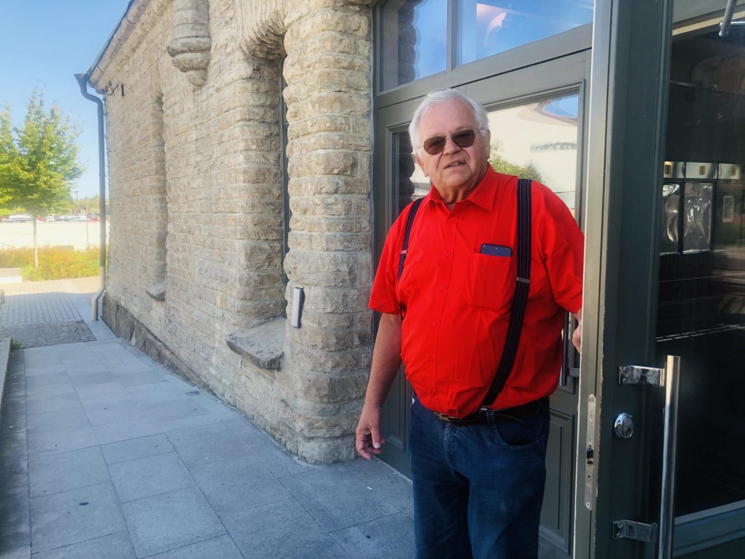 En äldre man i röd skjorta och hängslen står i en dörröppning till en stenbyggnad och tittar in i kameran.