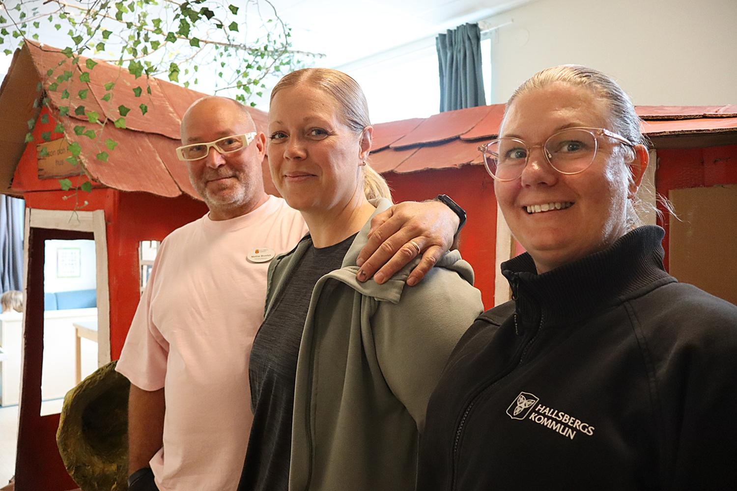 Tre vuxna står inomhus framför en liten röd lekstuga; två kvinnor bär glasögon och en man vilar sin hand på den ena kvinnans axel.