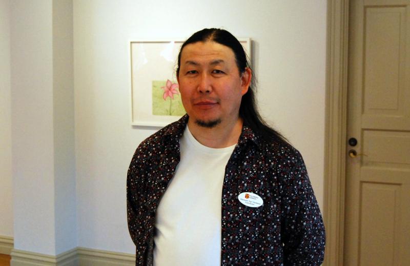 En man med långt mörkt hår står inomhus, klädd i en mönstrad skjorta över en vit t-shirt och en namnskylt. En inramad blomstertavla hänger på väggen bakom honom.