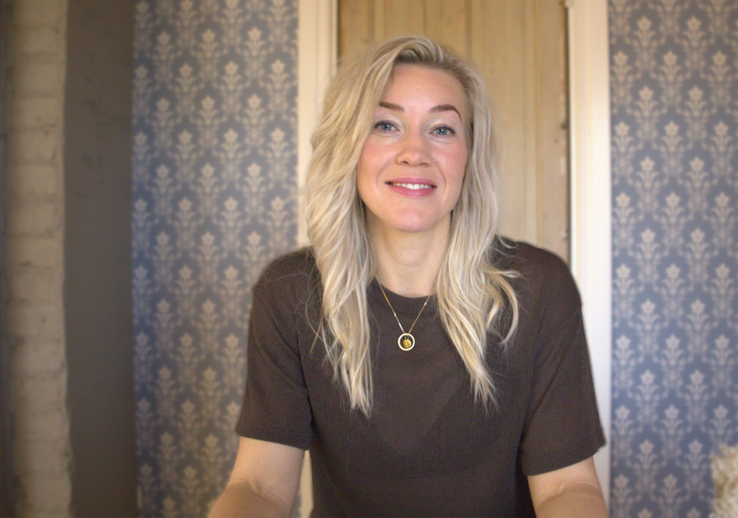 En kvinna med långt blont hår, klädd i en mörk topp och ett hängande halsband, sitter framför en mönstrad blå tapet och en trädörr och ler mot kameran.