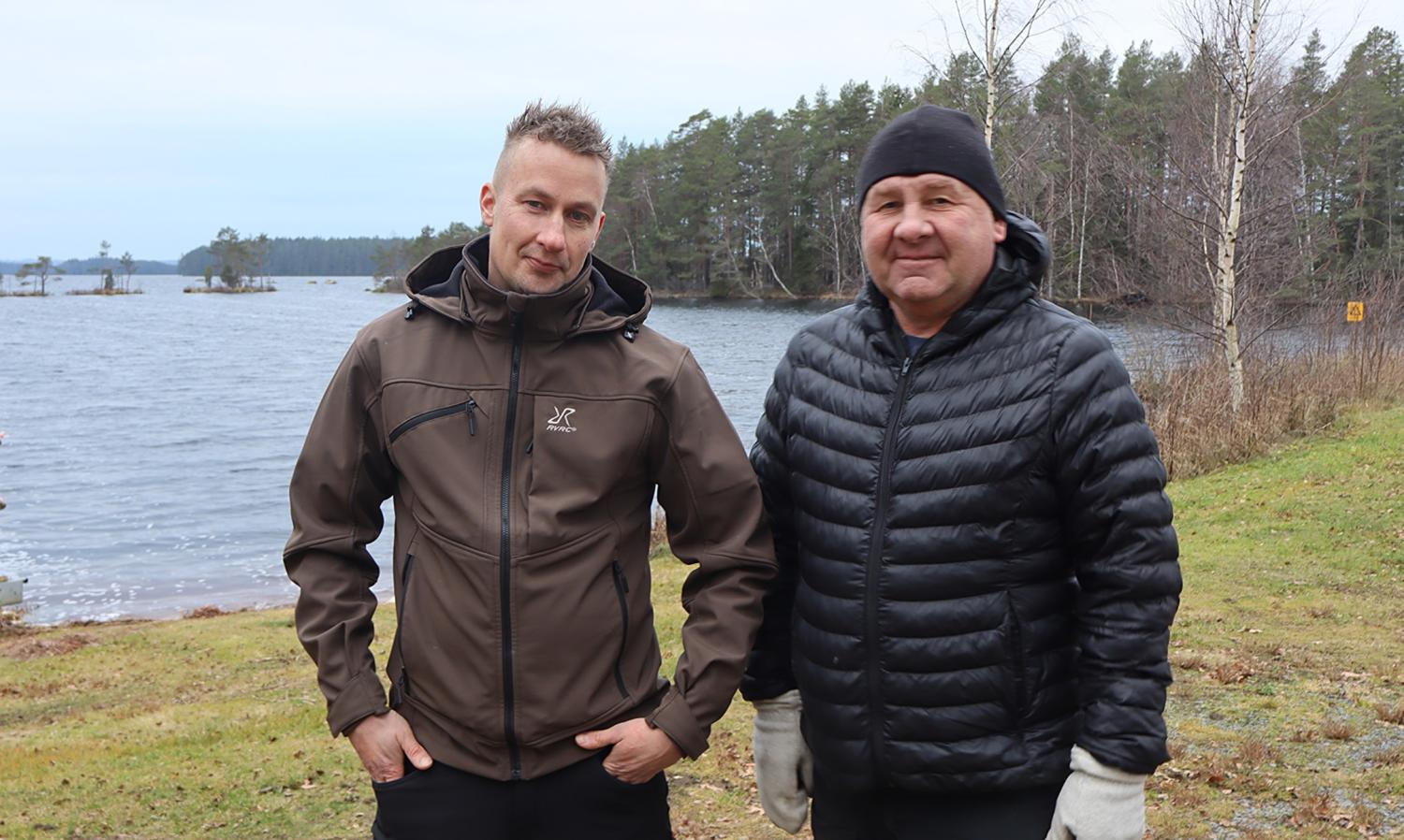 Två kavajklädda män står utomhus vid en sjö med träd och mulen himmel i bakgrunden.