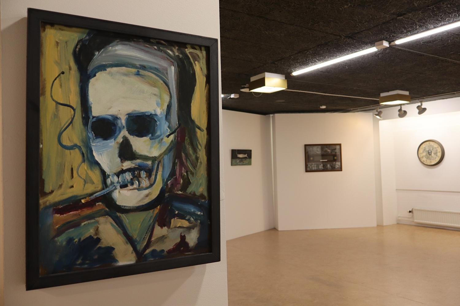 En målning av en rökande figur med dödskalleansikte visas på en gallerivägg, med ytterligare konstverk och en klocka i bakgrunden.