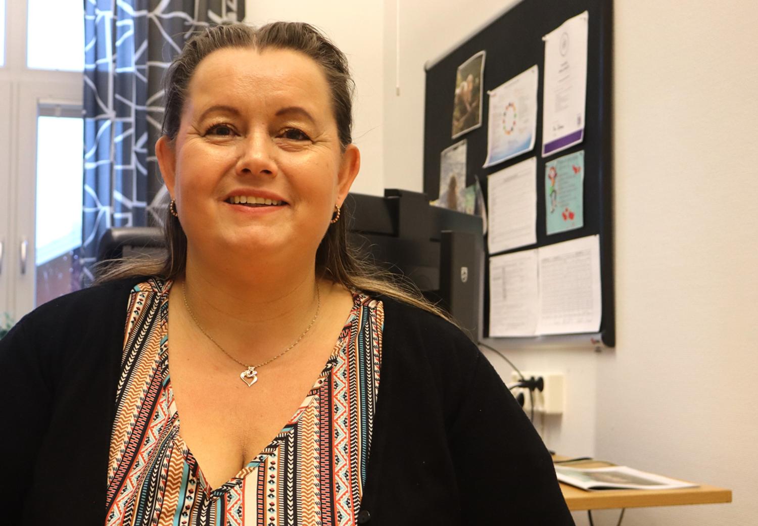 En kvinna med långt brunt hår och en mönstrad topp sitter och ler i en kontorsmiljö med en anslagstavla och papper i bakgrunden.