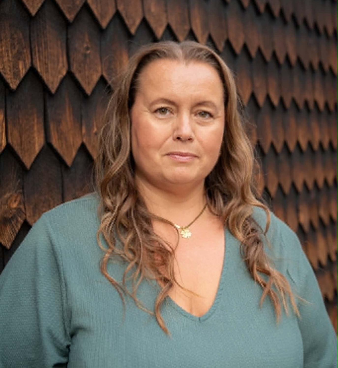 En kvinna med långt, vågigt brunt hår och en grön tröja står framför en vägg med mörka träpaneler.