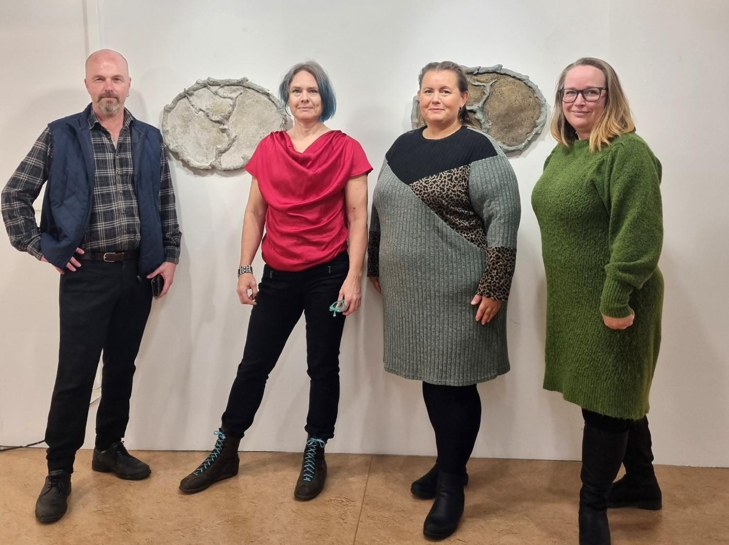 Fyra vuxna står sida vid sida inomhus framför en vit vägg med två abstrakta stenkonstverk. De är vända mot kameran och klädda i avslappnade vinterkläder.