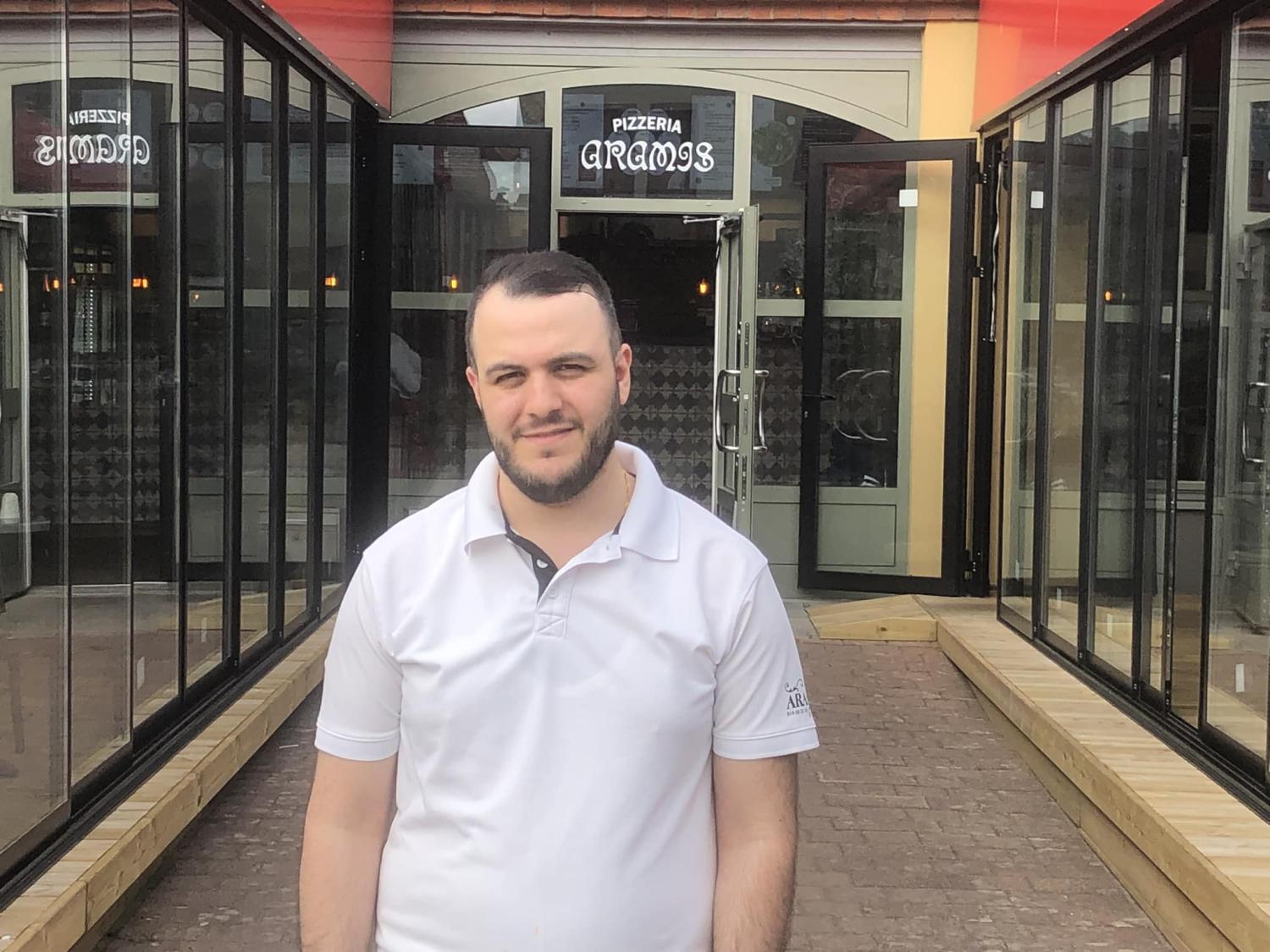 En man står framför en pizzeria med glasdörrar, klädd i en vit pikétröja. På skylten ovanför entrén står det 
