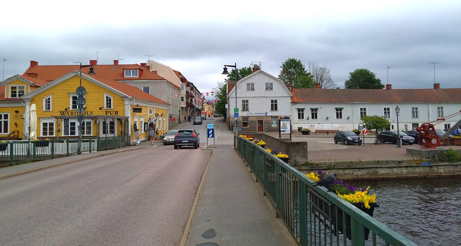Vy över en småstadsgata med en gul pub, vita byggnader, parkerade bilar och en bro som går över en kanal under en molnig himmel.