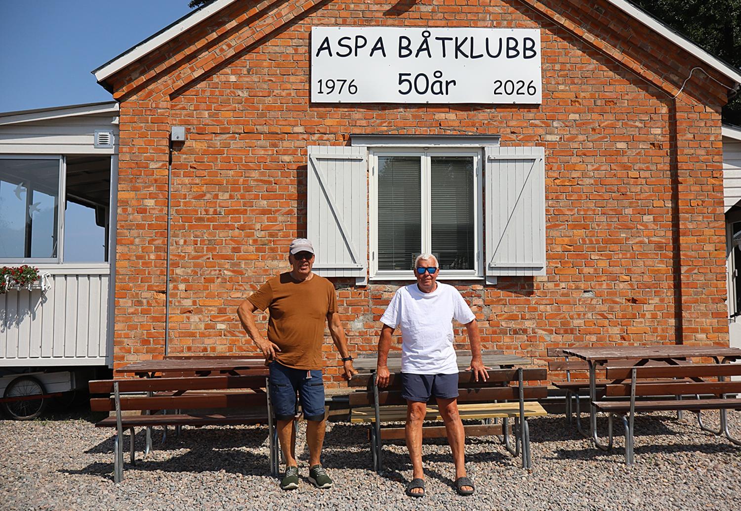 Två män står framför en tegelbyggnad med en skylt där det står ”Aspa Båtklubb 50 år 1976 2026”. Längs väggen står bänkar och på marken ligger grus.