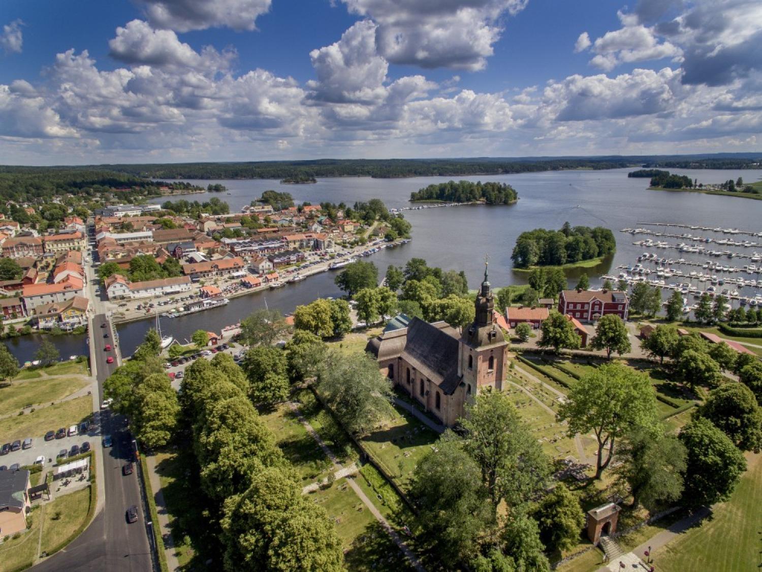 Flygfoto över en stad vid en sjö med en kyrka, marina, båtar och kluster av byggnader omgivna av gröna träd under en delvis molnig himmel.
