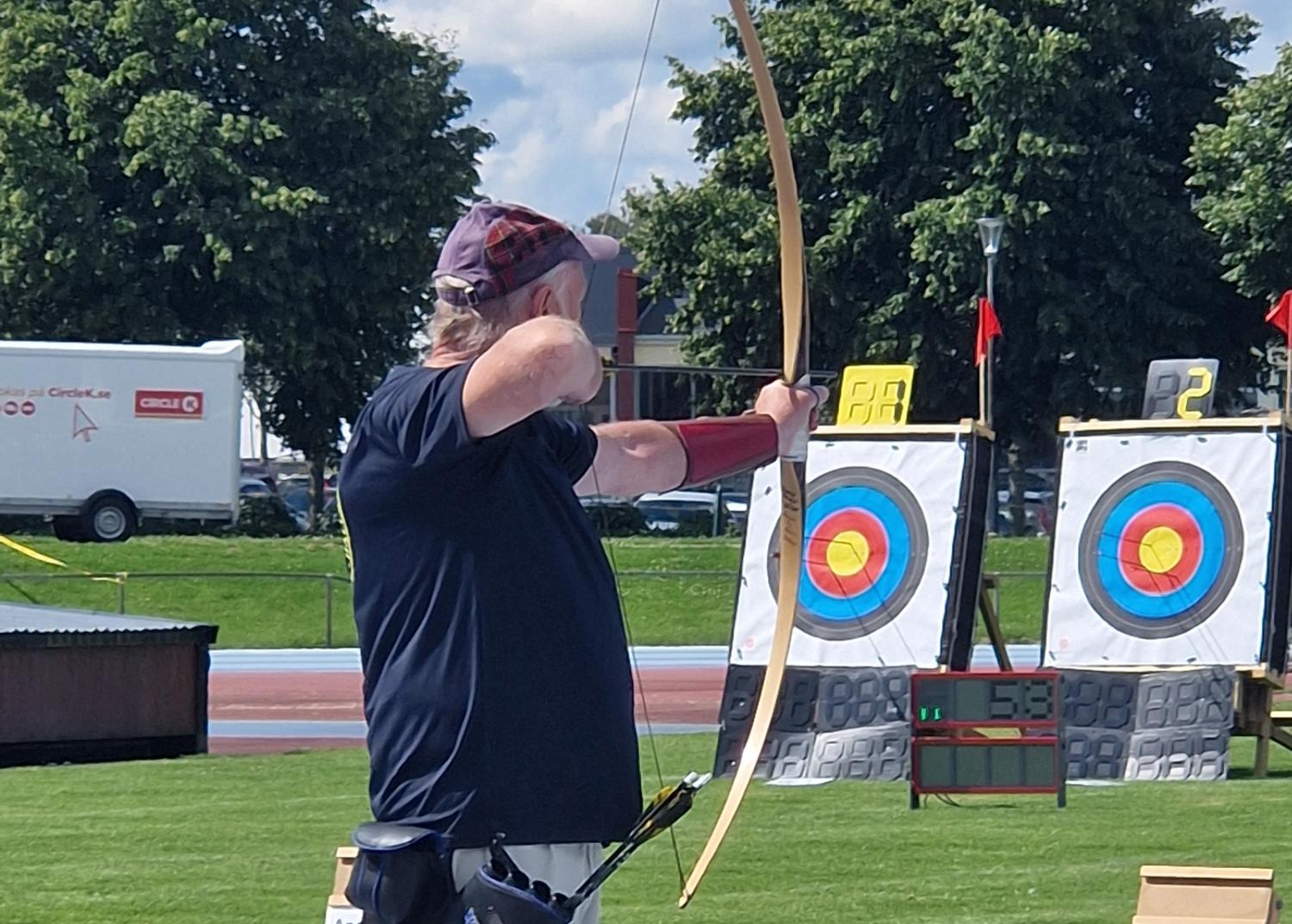 En person som bär keps och armskydd riktar en båge mot ett mål i en utomhus bågskyttebana en solig dag. Två cirkulära bågskyttemål är placerade i bakgrunden.