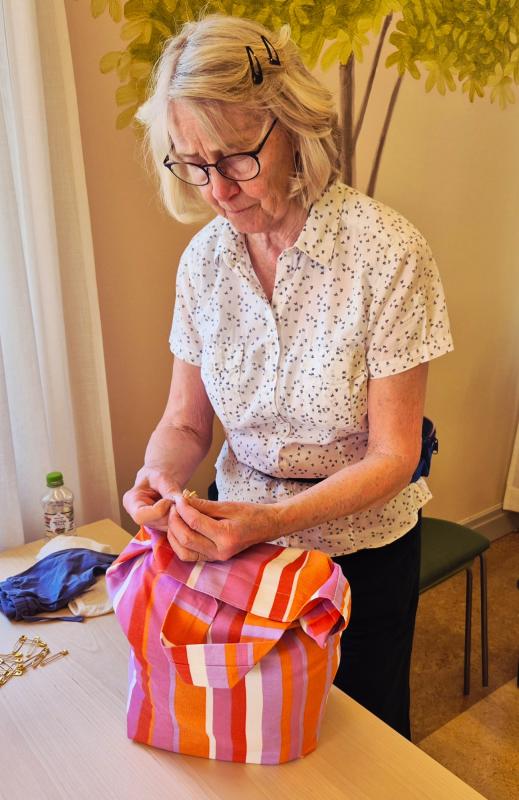 En äldre kvinna med glasögon viker ett papper medan hon står vid ett bord med en randig väska och några små föremål.