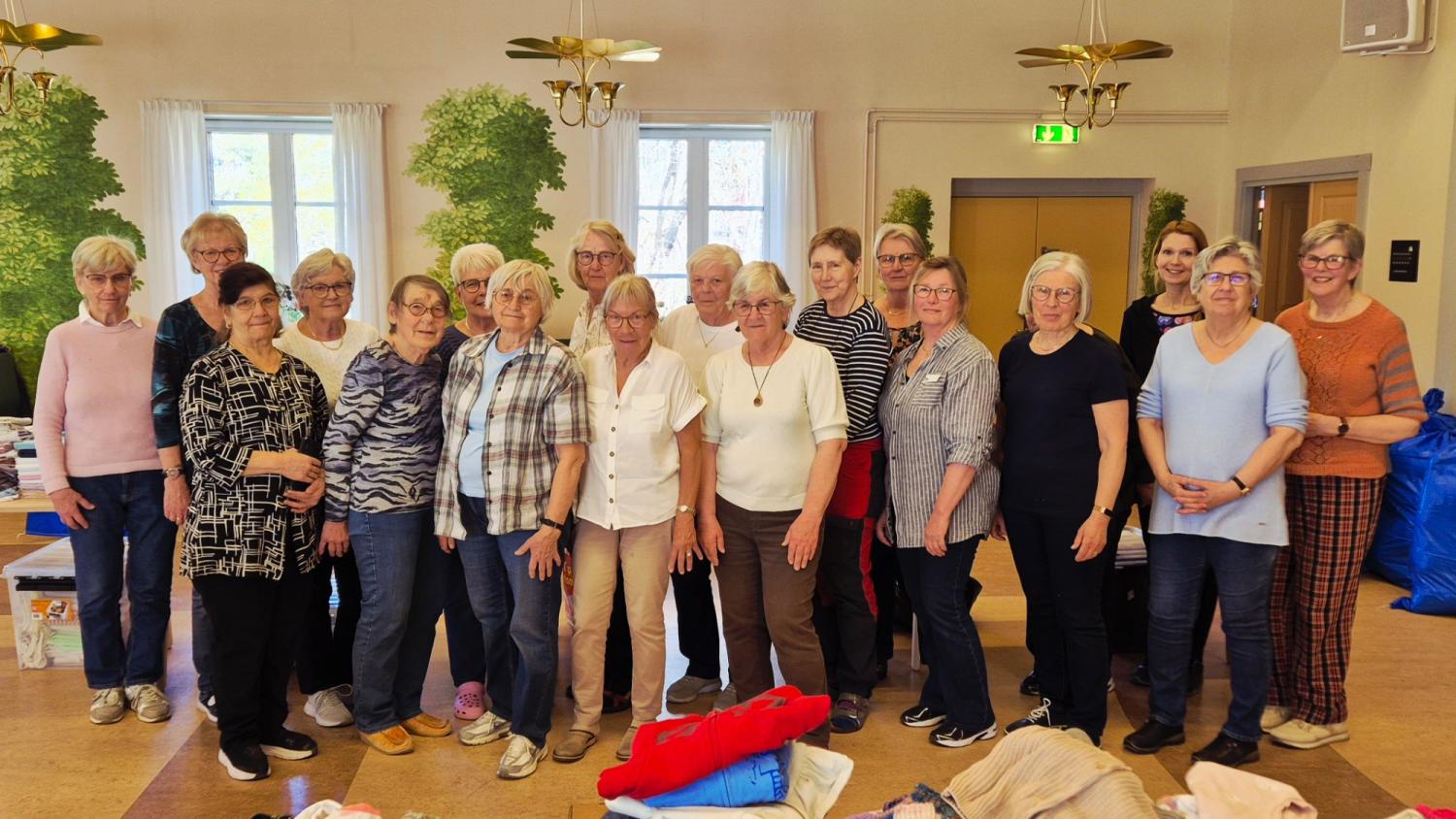 En grupp äldre kvinnor poserar tillsammans inomhus, stående i en linje. På borden framför dem syns hopvikta kläder och väskor.