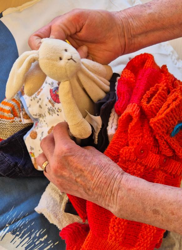 En persons händer sorterar i en hög med kläder, håller i en beige gosedjurskanin och arrangerar färgglada stickade plagg.