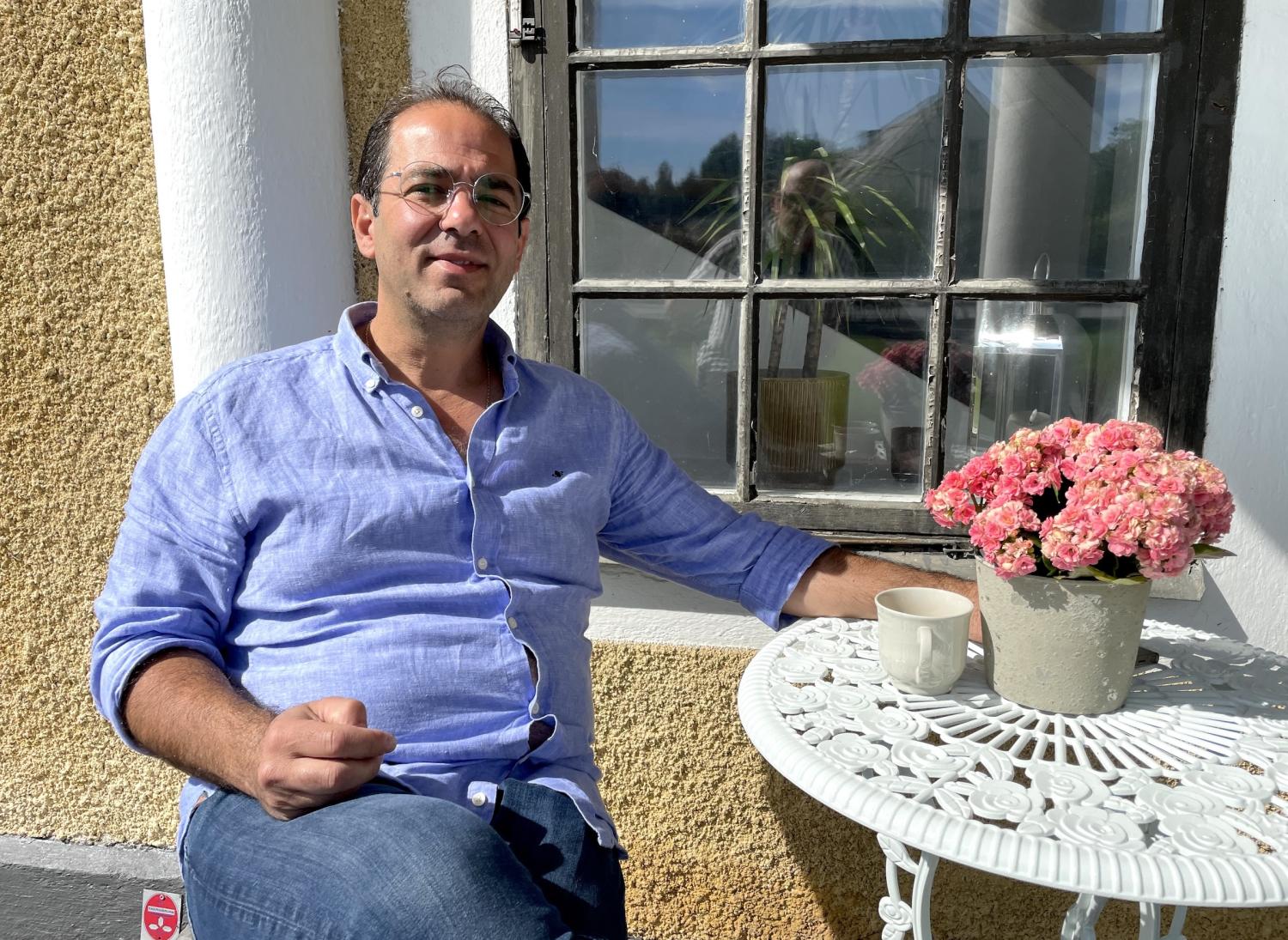 En man sitter vid ett vitt metallbord med rosa blommor och en mugg, ute vid ett fönster en solig dag.