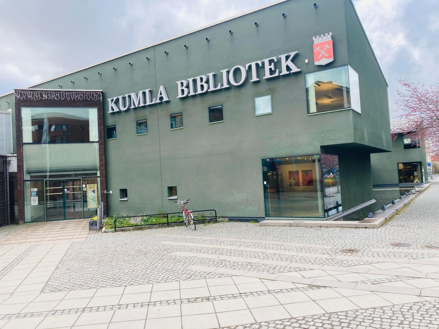 Exteriörvy av kumla bibliotek, en modern biblioteksbyggnad med gröna väggar och stora fönster, belägen på ett stenlagt torg med en cykel parkerad utanför.