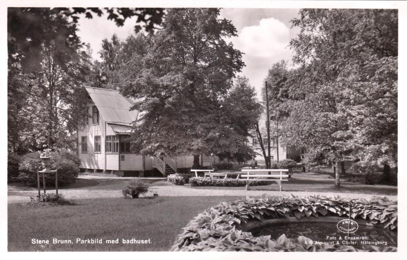 Svartvitt foto av ett trähus omgivet av träd, bänkar och en cirkelformad blomrabatt i en parkmiljö.