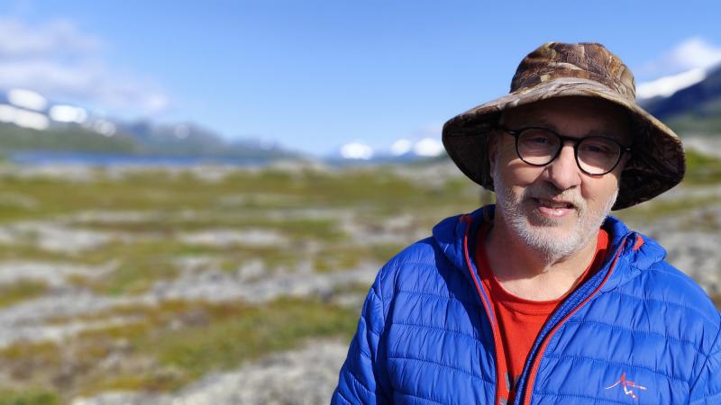 En man klädd i en blå jacka, röd skjorta, glasögon och en brun hatt står utomhus i ett soligt bergslandskap med snöfläckar i fjärran.