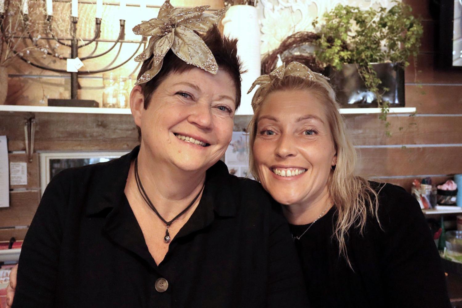 Två kvinnor med svarta toppar och pannband i bladguld ler mot varandra i ett varmt upplyst rum med växter och dekorationer i bakgrunden.