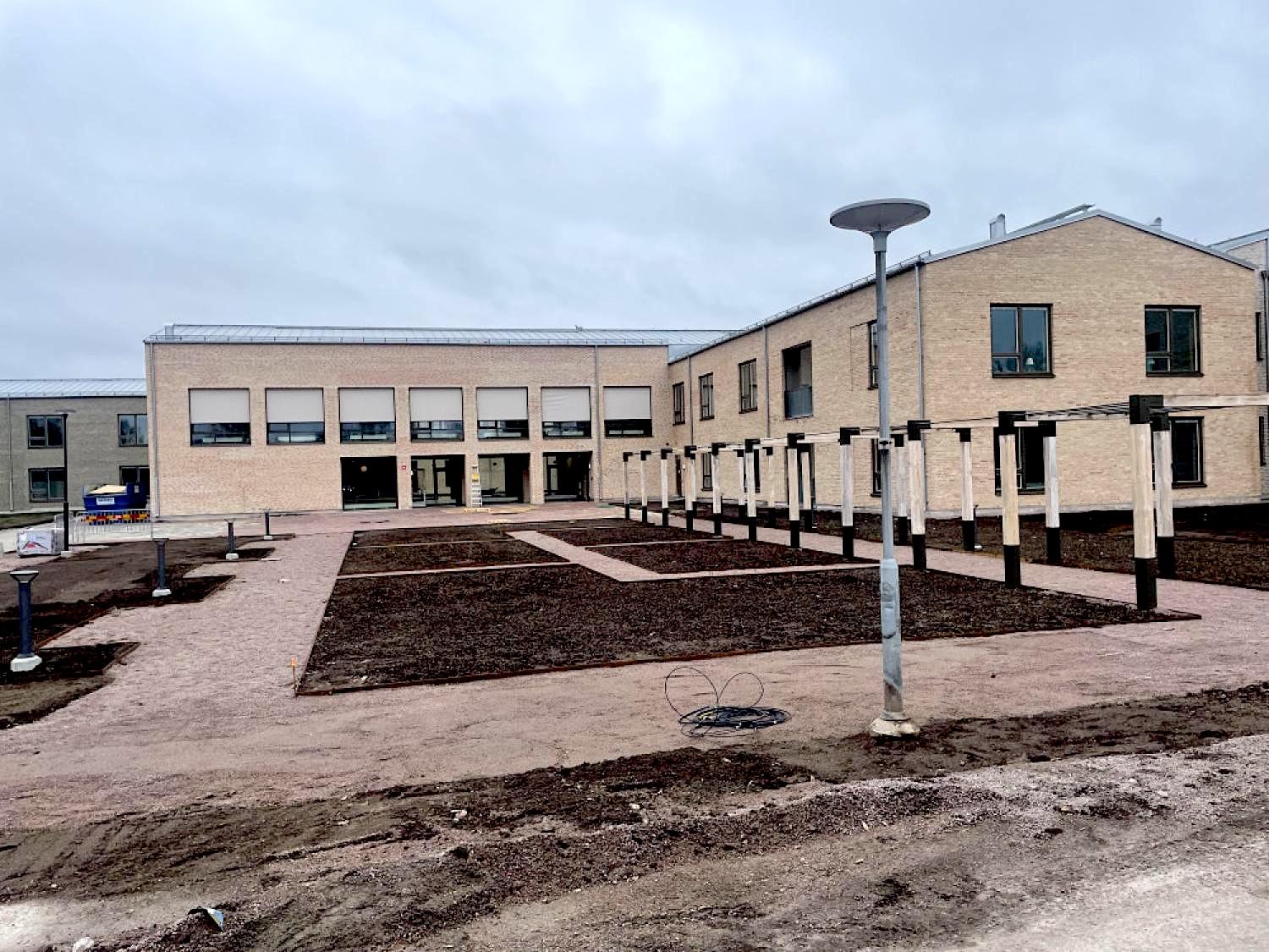 En modern tegelbyggnad med flera fönster och ett anlagt område med gångvägar och lyktstolpar framför, under en molnig himmel.