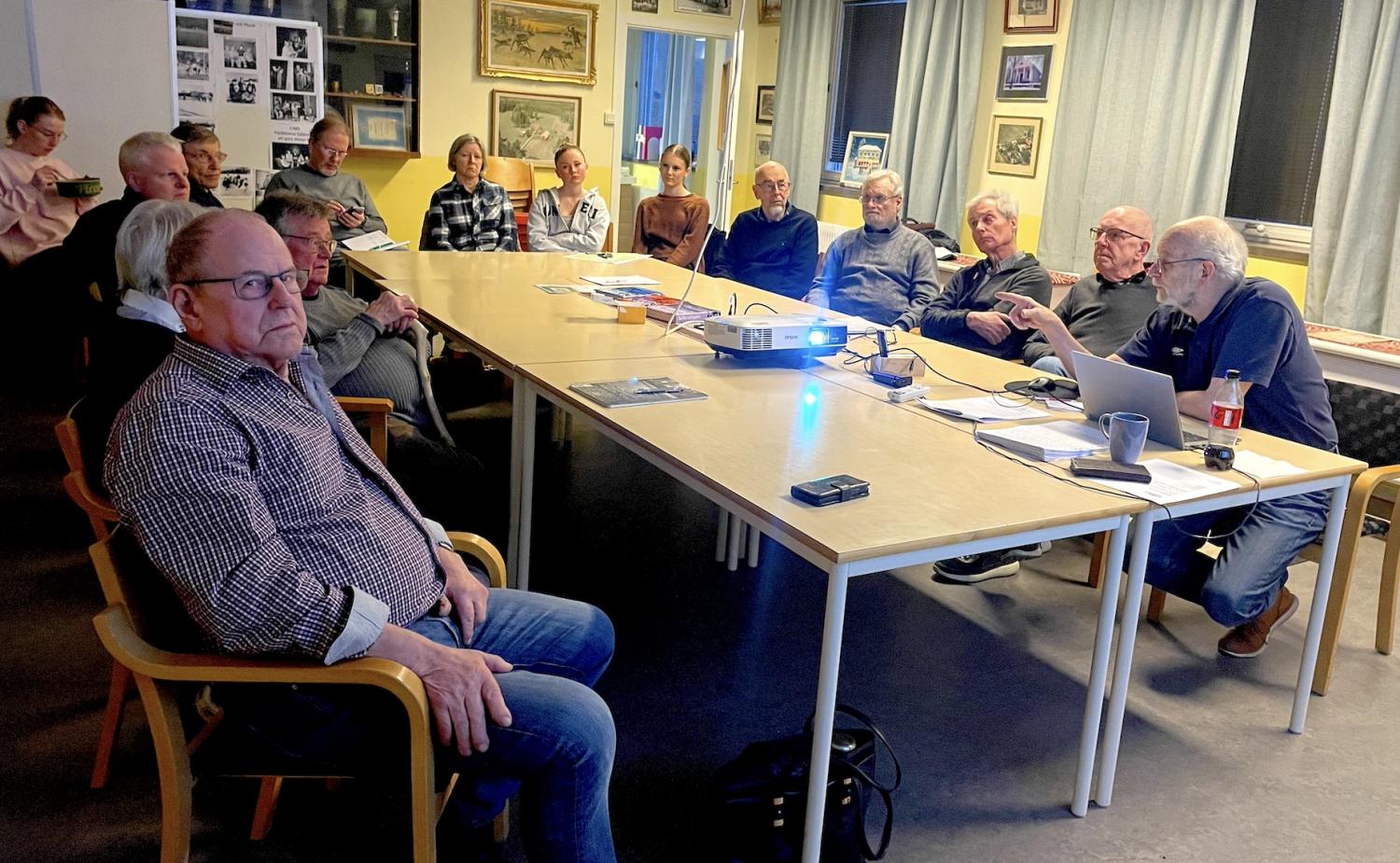 En grupp individer som uppmärksamt deltar i ett möte eller en presentation i ett rum med bilder på väggen.
