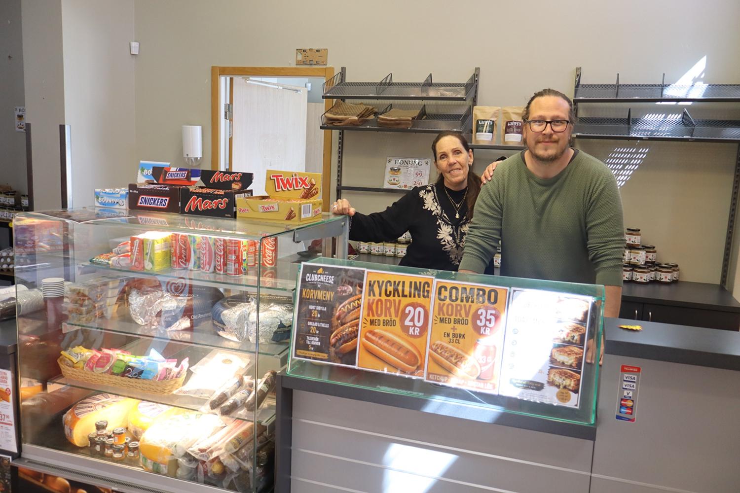 Två personer står bakom disken i en liten butik som säljer snacks, drycker och smörgåsar, med olika produkter utställda på hyllor och i glasmontrar.