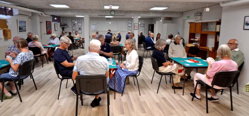 Flera grupper av äldre sitter vid bord i ett väl upplyst rum och spelar ett kortspel. Stämningen verkar lugn och koncentrerad.