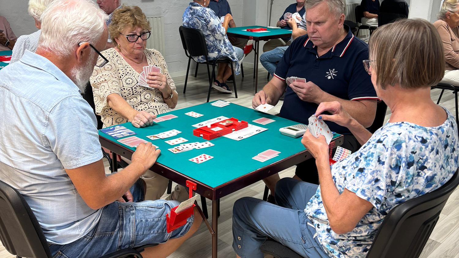 Fyra vuxna sitter runt ett grönt kortbord och spelar ett kortspel. I bakgrunden spelar andra grupper vid liknande bord.