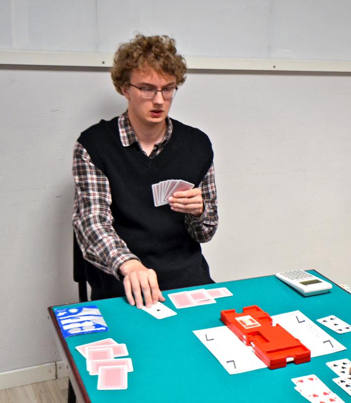 En person med glasögon och en rutig skjorta spelar ett kortspel vid ett bord där det finns kort, en miniräknare, en budlåda och ett blått etui.