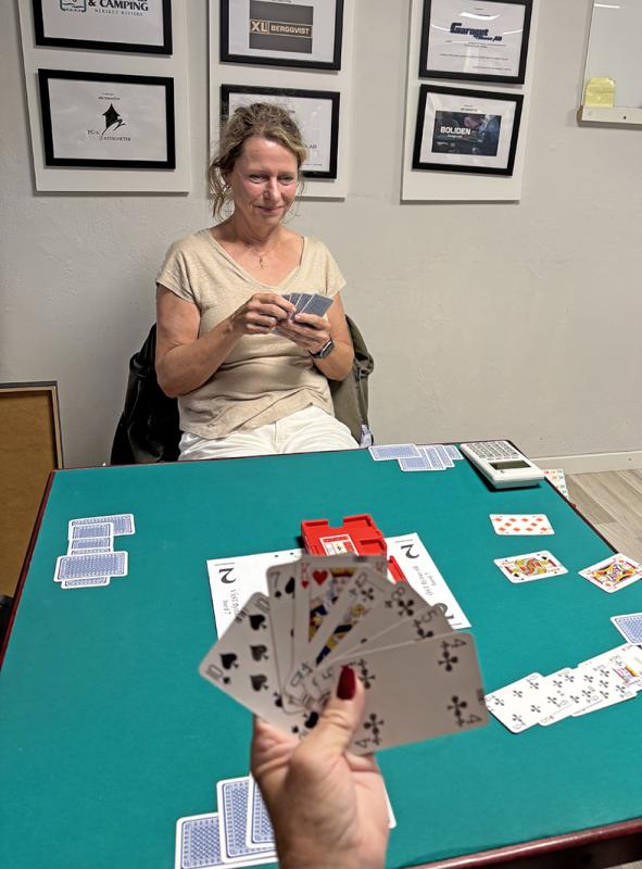 Två personer spelar ett kortspel vid ett grönt bord; den ena håller korten i handen och ler, medan den andras kort syns i förgrunden.