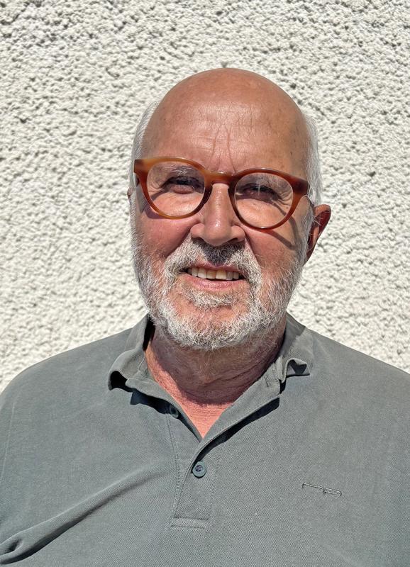 En äldre man med skalligt huvud, grått skägg och glasögon står utomhus i solskenet, klädd i en grå polotröja framför en strukturerad vit vägg.