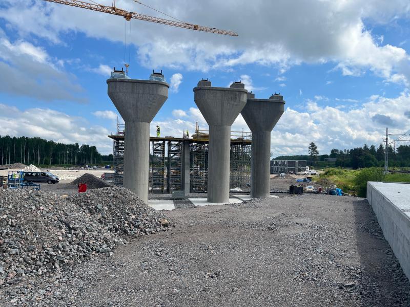 Fyra stora bropelare av betong under uppförande på en byggarbetsplats där byggnadsställningar, maskinutrustning och en kran syns mot en delvis molnig himmel.