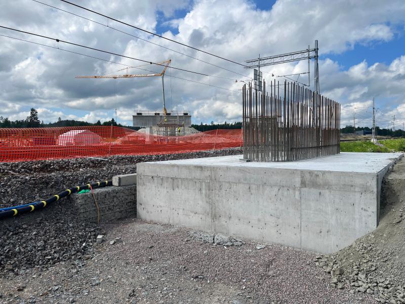 Betongfundament med vertikala armeringsjärn på en järnvägsbyggarbetsplats, orange säkerhetsstängsel, kran och molnig himmel i bakgrunden.