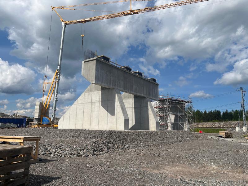 En stor bärkonstruktion för en betongbro under uppförande, med kranar och byggnadsställningar i närheten, på en grusplan under en delvis molnig himmel.