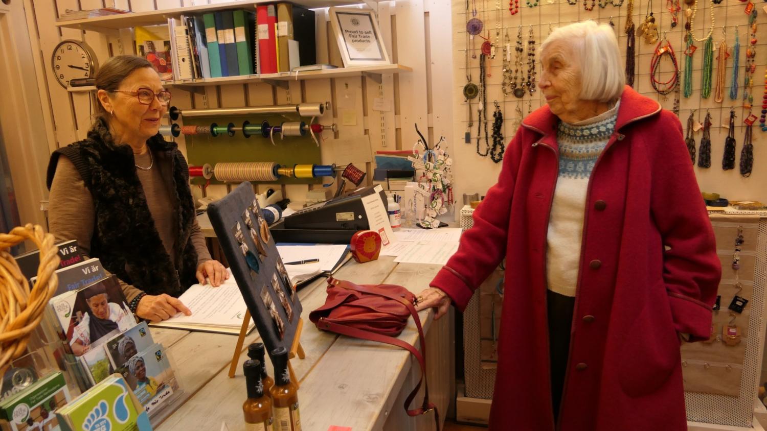 Två äldre kvinnor samtalar vid en butiksdisk; den ena står bakom disken, den andra i röd rock står framför, med hantverk och förnödenheter synliga i bakgrunden.