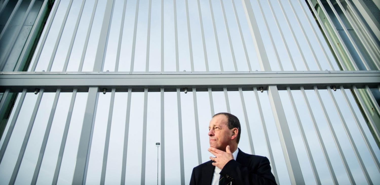 En kostymklädd man står framför ett högt metallstaket eller en grind och tittar åt sidan med handen nära hakan.