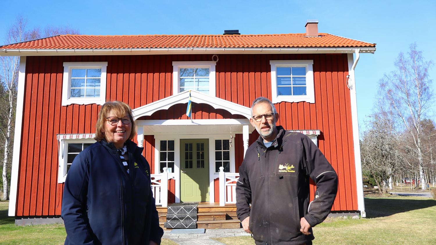 Två personer står framför ett rött trähus med vit dekor en solig dag.