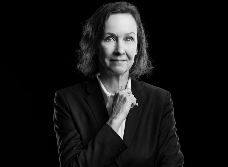 En kvinna i mörk kostym poserar mot en svart bakgrund och tittar in i kameran med ett neutralt uttryck och handen vilande nära hakan.