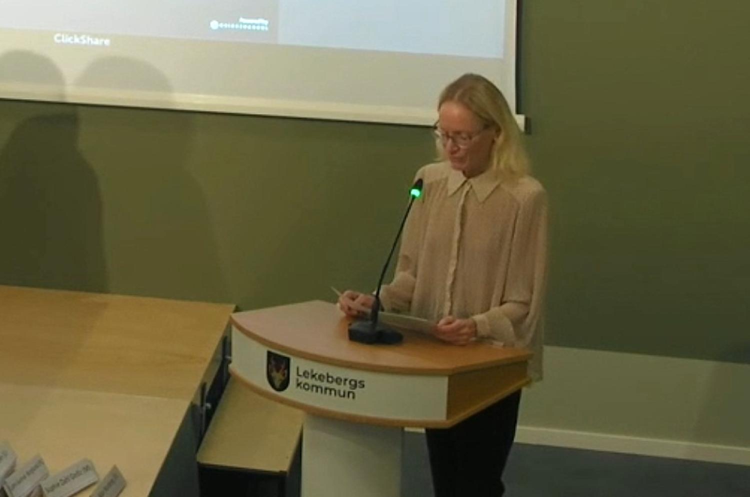 En person står vid ett talarstativ med texten ”Lekebergs kommun” och talar i en mikrofon i ett konferensrum.