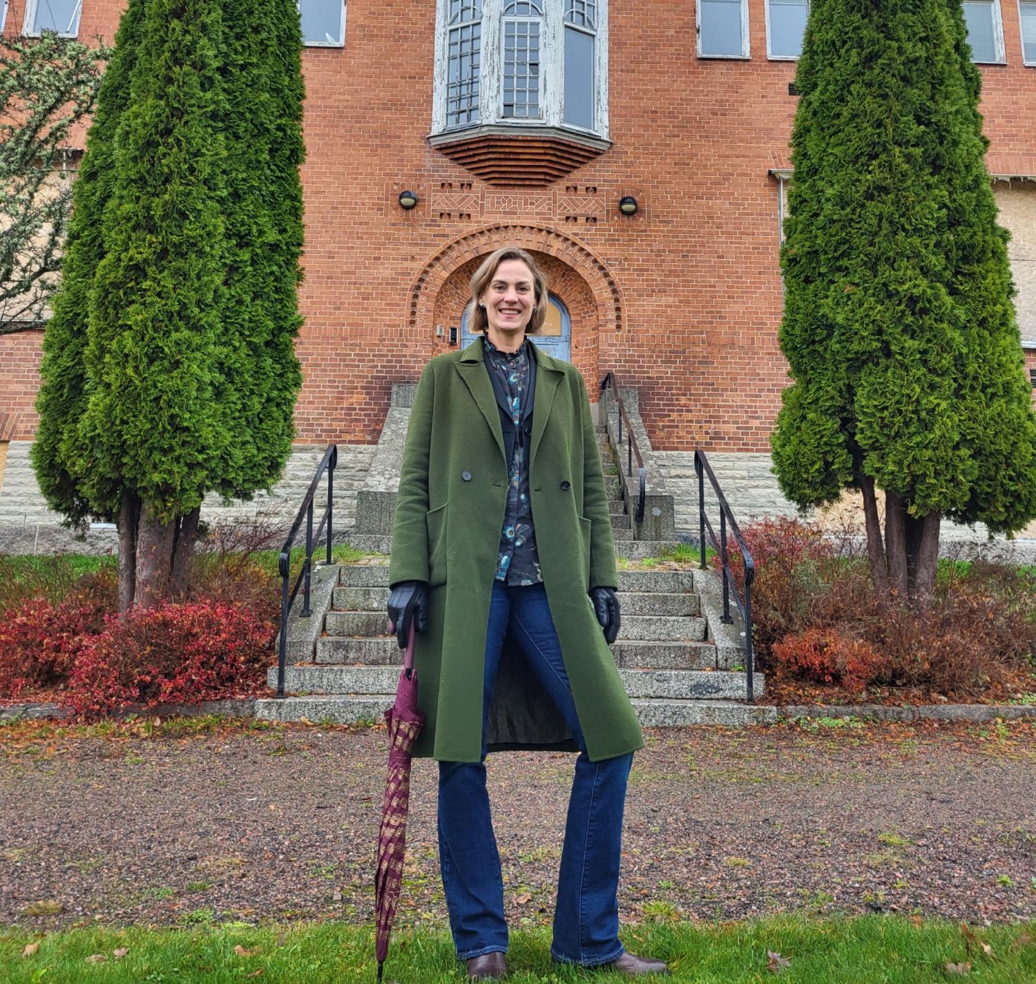 En person i grön kappa står på en stentrappa framför en tegelbyggnad, med ett stängt paraply och flankerad av två höga träd.