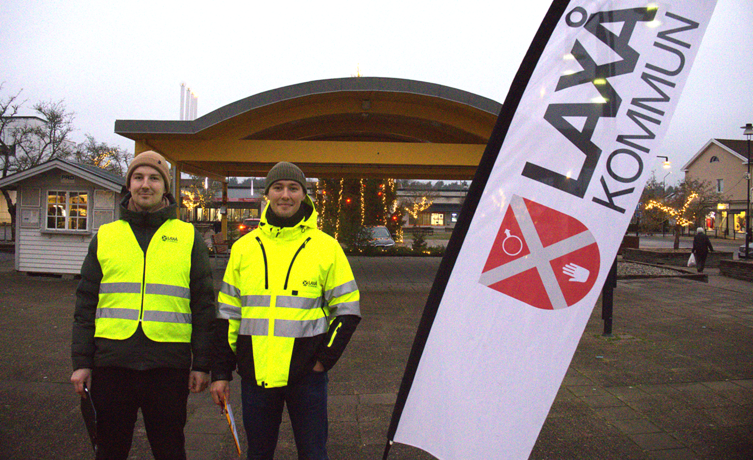 Två personer i gula reflexvästar står bredvid en flagga med texten 