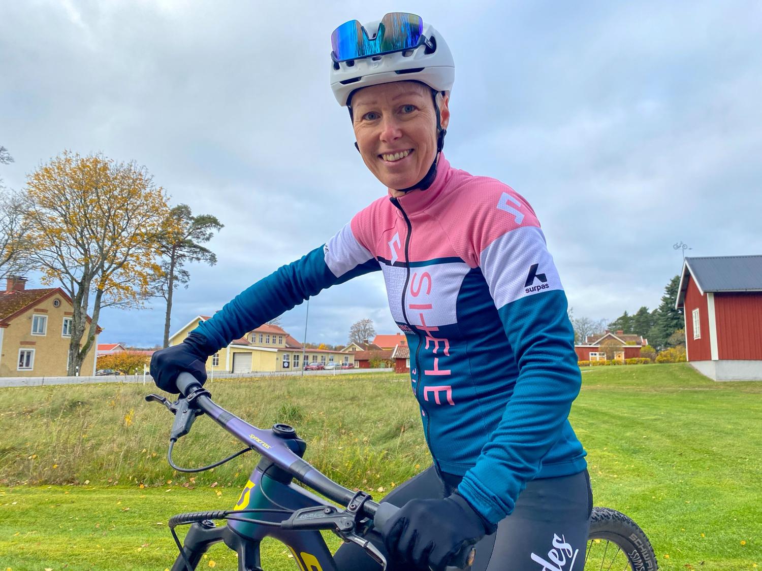 En person i cykelkläder och hjälm ler mot kameran medan han håller i en cykel utomhus i ett bostadsområde.