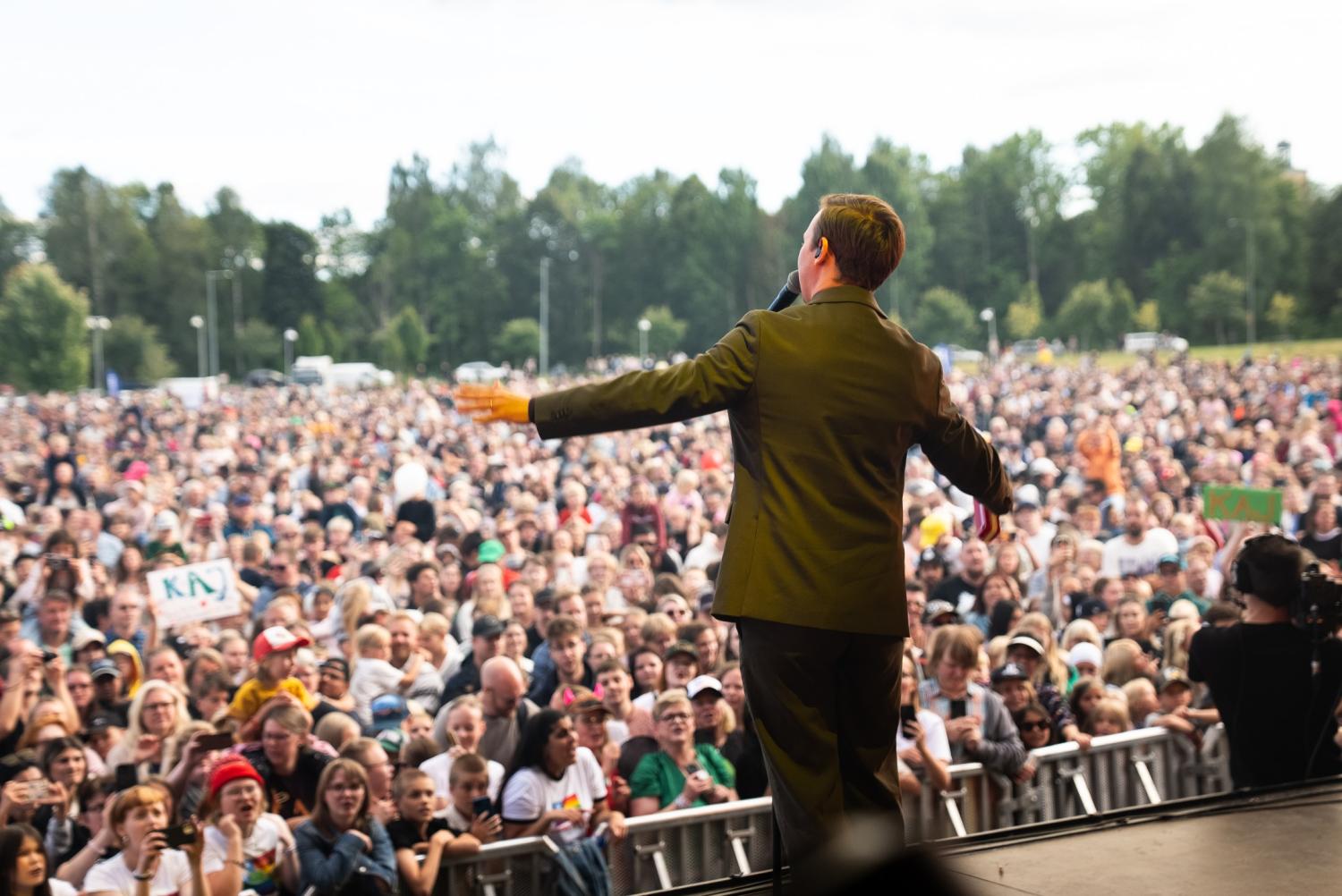 En person i kostym talar eller uppträder på en utomhusscen inför en stor publik, med träd och himmel i bakgrunden.