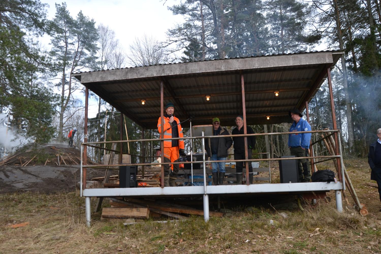 Fyra personer står på en täckt utomhusplattform omgiven av träd, och en person i ljusorangea arbetskläder talar till de andra.